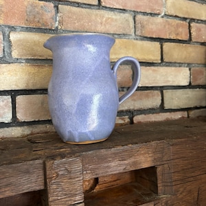 May include: A handmade, light purple ceramic pitcher with a handle and a pouring spout. The pitcher has a smooth, glossy finish and sits on a wooden surface. The background features a brick wall.