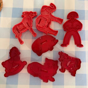 May include: A set of seven red plastic cookie cutters in various shapes, including an elephant, a donkey, a person, a pig, a rocking horse, a cat, and a dog. Each cutter features molded details on the surface and a raised edge for cutting dough.
