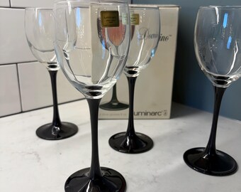 May include: A set of four clear glass wine glasses with black stems and bases. Each glass features a rounded bowl and a slender black stem. The original cardboard packaging box is visible in the background, labeled with the text Luminarc Domino.