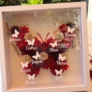 Peut inclure: Un cadre avec une œuvre d'art en forme de papillon floral. Le papillon est composé de fleurs en papier rouge, or et bordeaux. Des papillons blancs et des noms sont placés dans la conception. Le cadre est blanc et porte les mots "Maman" et "Marie" en lettres dorées.