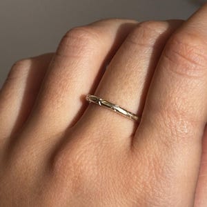 May include: A slender gold ring with a textured pattern is displayed on a finger. The ring's detailed design reflects light, highlighting its fine craftsmanship. The ring is a simple yet elegant piece of jewellery.