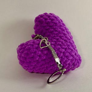 May include: A vibrant purple, heart-shaped item made from a plush material, with a silver-coloured chain and key ring. The heart has a textured surface. A small heart charm is attached.