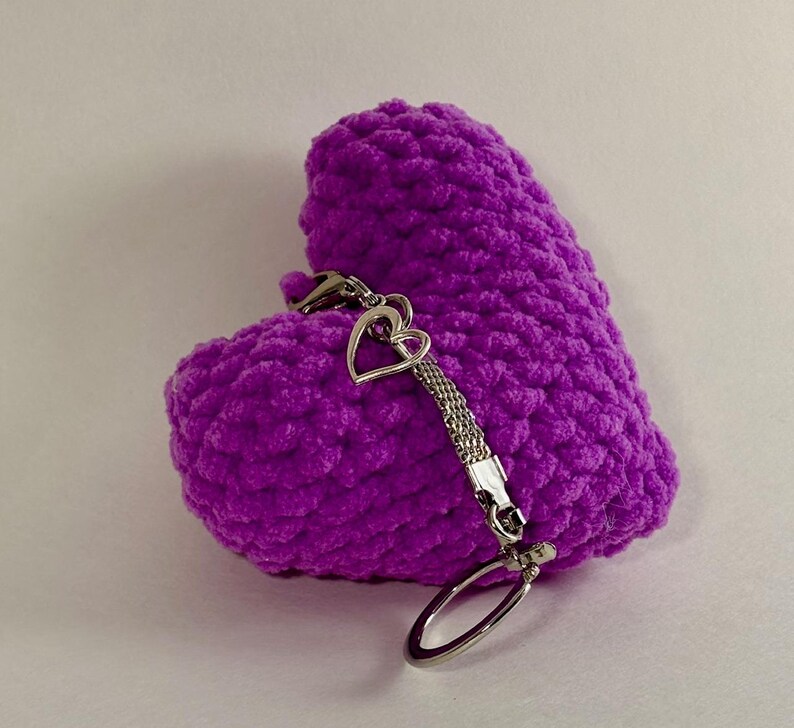 May include: A vibrant purple, heart-shaped item made from a plush material, with a silver-coloured chain and key ring. The heart has a textured surface. A small heart charm is attached.