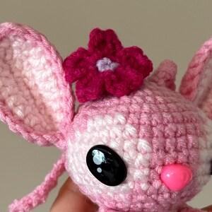 Op de afbeelding: Een handgemaakte gehaakte pop in roze en witte tinten, met grote zwarte ogen en een roze neus. Een magenta bloem siert de bovenkant van het hoofd van de pop. De oren van de pop zijn groot en afgerond.