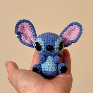 Op de afbeelding: Een handgemaakte blauwe en lichtblauwe gehaakte Stitch-pop met grote zwarte ogen en oversized oren. De oren hebben een roze binnenkant en blauwe rand. De pop wordt in een hand gehouden.