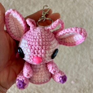 Op de afbeelding: Handgemaakte roze gehaakte sleutelhanger met een stripfiguur met grote zwarte ogen, een roze neus en paarse accenten op de voeten. Het heeft een zilveren sluiting. De oren zijn groot en slap.