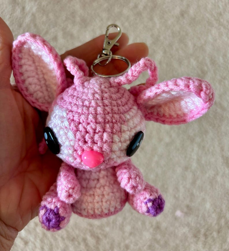 Op de afbeelding: Handgemaakte roze gehaakte sleutelhanger met een stripfiguur met grote zwarte ogen, een roze neus en paarse accenten op de voeten. Het heeft een zilveren sluiting. De oren zijn groot en slap.