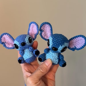 Op de afbeelding: Twee handgemaakte gehaakte figuren van een blauw alien-personage met grote oren en zwarte ogen. De figuren zijn blauw met lichtblauwe en roze accenten. De figuren worden in de hand van een persoon gehouden.