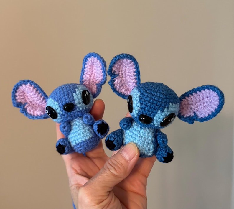 Op de afbeelding: Twee handgemaakte gehaakte figuren van een blauw alien-personage met grote oren en zwarte ogen. De figuren zijn blauw met lichtblauwe en roze accenten. De figuren worden in de hand van een persoon gehouden.