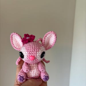 Op de afbeelding: Een handgemaakte gehaakte pop in roze en wit, met grote zwarte ogen en een roze neus. De pop heeft een bloem op haar hoofd en paarse accenten op de voeten. Dit schattige knuffel is perfect voor kinderen of verzamelaars.