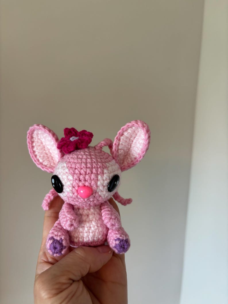 Op de afbeelding: Een handgemaakte gehaakte pop in roze en wit, met grote zwarte ogen en een roze neus. De pop heeft een bloem op haar hoofd en paarse accenten op de voeten. Dit schattige knuffel is perfect voor kinderen of verzamelaars.