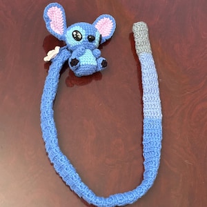 Op de afbeelding: Een handgemaakte blauwe Stitch-gehaakte pop met grote oren, zwarte ogen en een kleine witte bloem. De pop is bevestigd aan een lang, blauw, verlopend gehaakt koord, perfect voor een kinderspeelgoed of een uniek accessoire.