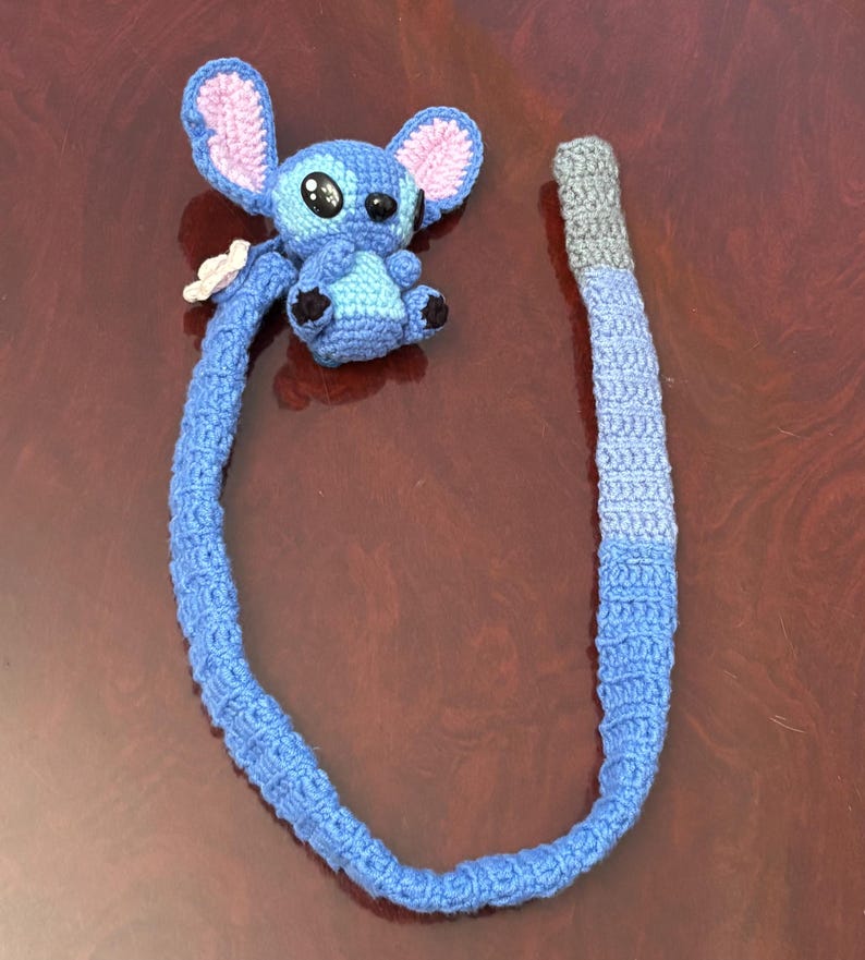 Op de afbeelding: Een handgemaakte blauwe Stitch-gehaakte pop met grote oren, zwarte ogen en een kleine witte bloem. De pop is bevestigd aan een lang, blauw, verlopend gehaakt koord, perfect voor een kinderspeelgoed of een uniek accessoire.