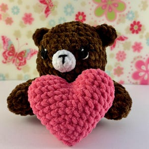 May include: A brown teddy bear holding a pink heart. The bear has a white muzzle with a black "T" shape and black eyes. The heart is a vibrant pink colour, and the background has a floral pattern.