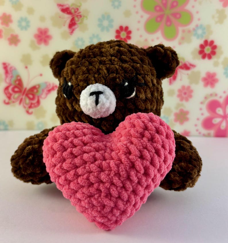May include: A brown teddy bear holding a pink heart. The bear has a white muzzle with a black "T" shape and black eyes. The heart is a vibrant pink colour, and the background has a floral pattern.