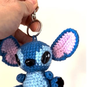 Op de afbeelding: Gehaakte Stitch sleutelhanger in blauw en roze. Het handgemaakte pluche speelgoed heeft grote zwarte ogen en oversized oren. De sleutelhanger is bevestigd aan een zilveren metalen ring en sluiting. Een schattig en uniek accessoire.