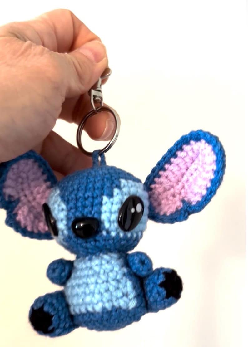 Op de afbeelding: Gehaakte Stitch sleutelhanger in blauw en roze. Het handgemaakte pluche speelgoed heeft grote zwarte ogen en oversized oren. De sleutelhanger is bevestigd aan een zilveren metalen ring en sluiting. Een schattig en uniek accessoire.