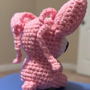 Op de afbeelding: Een handgemaakte roze gehaakte konijnenpop met lange, slappe oren en een kleine staart. Het amigurumi-konijn heeft zwarte ogen en een witte snuit. De pop is gemaakt met een getextureerde steek, waardoor hij een zacht uiterlijk heeft.