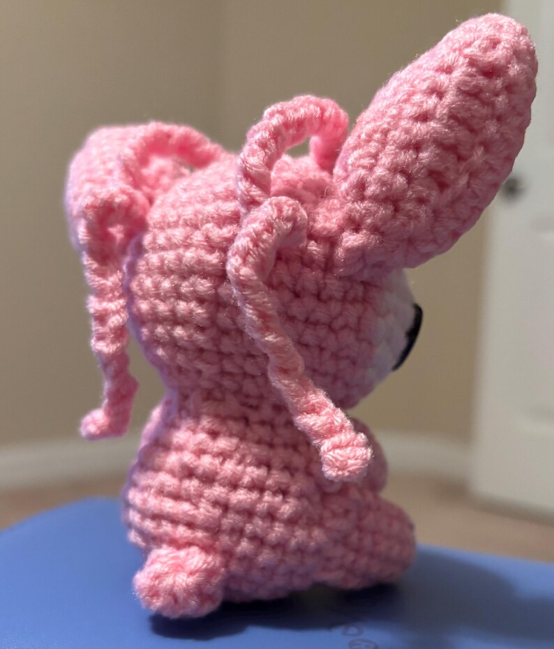 Op de afbeelding: Een handgemaakte roze gehaakte konijnenpop met lange, slappe oren en een kleine staart. Het amigurumi-konijn heeft zwarte ogen en een witte snuit. De pop is gemaakt met een getextureerde steek, waardoor hij een zacht uiterlijk heeft.
