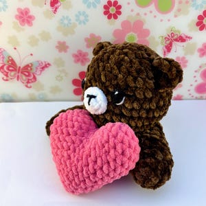 May include: A brown crochet bear holding a pink heart. The bear has a white muzzle and black eyes. The heart is a vibrant pink colour, and the bear is made of soft, textured yarn. The background has a floral pattern.