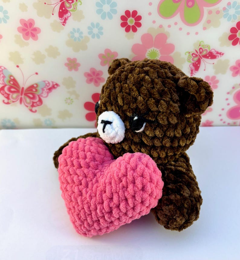 May include: A brown crochet bear holding a pink heart. The bear has a white muzzle and black eyes. The heart is a vibrant pink colour, and the bear is made of soft, textured yarn. The background has a floral pattern.