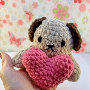 May include: A handmade crochet dog with brown ears and a beige body, holding a pink heart. The dog has black eyes and a stitched mouth. The plush toy is soft and cuddly, ideal for a child or as a decorative item.
