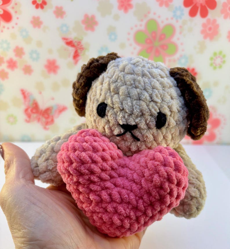 May include: A handmade crochet dog with brown ears and a beige body, holding a pink heart. The dog has black eyes and a stitched mouth. The plush toy is soft and cuddly, ideal for a child or as a decorative item.