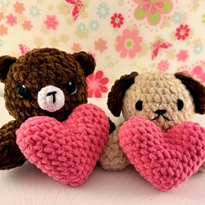 May include: Two handmade crochet toys: a brown bear and a tan dog, each holding a pink heart. The bear has a white muzzle and black eyes. The dog has brown ears and a black nose. The background features a floral pattern.