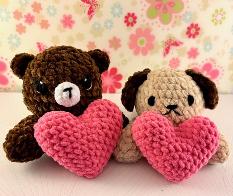 May include: Two handmade crochet toys: a brown bear and a tan dog, each holding a pink heart. The bear has a white muzzle and black eyes. The dog has brown ears and a black nose. The background features a floral pattern.