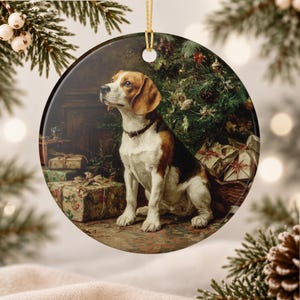 Puede incluir: Un adorno de cerámica redondo con una ilustración detallada de un perro beagle sentado frente a un árbol de Navidad decorado con regalos envueltos. El perro tiene pelaje marrón, negro y blanco y lleva un collar oscuro. Un hilo dorado cuelga arriba.