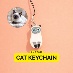 May include: A custom cat keychain featuring a Siamese cat charm with blue eyes, a cream-coloured body, and dark brown ears and paws. The charm hangs from a black cord. A circular inset shows a photograph of a real Siamese cat. Text reads CUSTOM CAT KEYCHAIN.