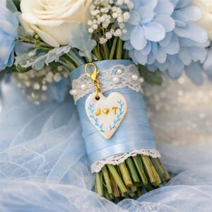 Op de afbeelding: Een boeket van witte rozen en lichtblauwe bloemen omwikkeld met lichtblauw lint en witte kant. Een hartvormige bedel met de letters J en T en een klein gouden hartje hangt aan een gouden sluiting bevestigd aan het handvat van het boeket.