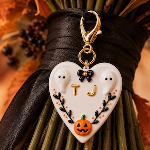 May include: A white heart-shaped charm featuring two white ghosts, a black bow, the gold letters T and J, and an orange jack-o'-lantern at the bottom. The charm is accented with black leafy vines and small orange dots, attached to a gold lobster clasp.