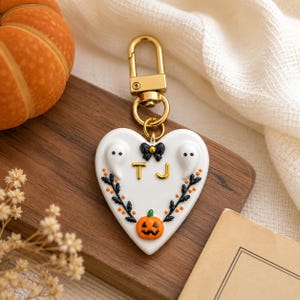 May include: A white heart-shaped keychain featuring a gold-tone metal lobster clasp. The design includes two small white ghosts, a black bow, a small orange pumpkin, and black leafy branches with orange dots. The gold letters T and J are centered.