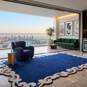 May include: A modern living room with a blue rug featuring a white decorative border. A blue armchair and green sofa are visible. The room has a view of a cityscape and a balcony.