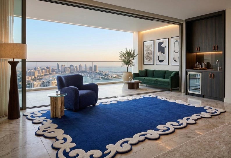 May include: A modern living room with a blue rug featuring a white decorative border. A blue armchair and green sofa are visible. The room has a view of a cityscape and a balcony.