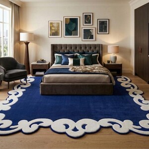 May include: A bedroom with a large, dark blue rug with a white, scalloped border. The room features a bed with a tufted headboard, side tables with lamps, and artwork on the wall. A dark gray armchair sits near a window.