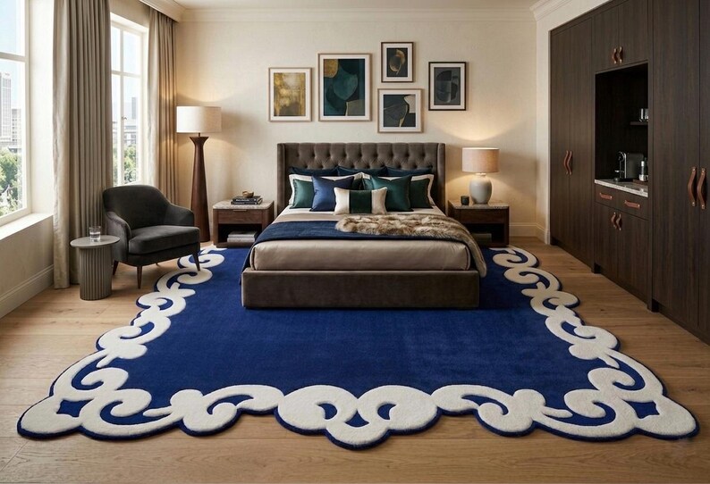 May include: A bedroom with a large, dark blue rug with a white, scalloped border. The room features a bed with a tufted headboard, side tables with lamps, and artwork on the wall. A dark gray armchair sits near a window.