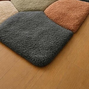 May include: Close-up of a multi-colored rug with a geometric pattern. The rug features square sections in shades of gray, beige, green, and orange. The rug has a soft, textured appearance, and is placed on a wooden floor.