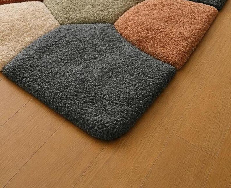 May include: Close-up of a multi-colored rug with a geometric pattern. The rug features square sections in shades of gray, beige, green, and orange. The rug has a soft, textured appearance, and is placed on a wooden floor.