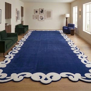 May include: A large, rectangular blue rug with a white, ornate border. The rug is the focal point of a room with hardwood floors, green sofas, and framed artwork. The rug's dimensions are approximately 10 x 15 feet.