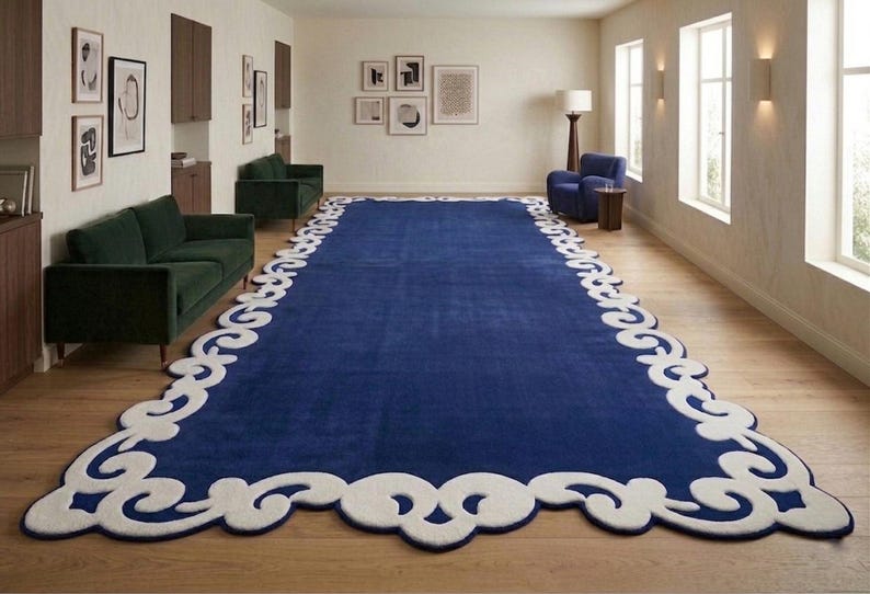 May include: A large, rectangular blue rug with a white, ornate border. The rug is the focal point of a room with hardwood floors, green sofas, and framed artwork. The rug's dimensions are approximately 10 x 15 feet.