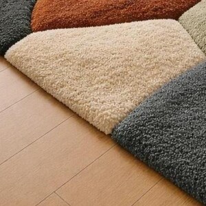 May include: Close-up of a multi-colored rug with a geometric pattern. The rug features sections of gray, beige, orange, and tan. The rug is placed on a light brown wooden floor.