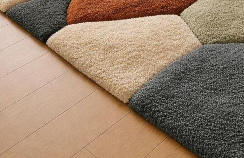 May include: Close-up of a multi-colored rug with a geometric pattern. The rug features sections of gray, beige, orange, and tan. The rug is placed on a light brown wooden floor.