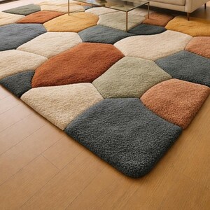 May include: A large, textured area rug with a geometric pattern. The rug features irregular, stone-like shapes in various colors, including beige, gray, orange, and yellow. The rug is placed on a wooden floor.