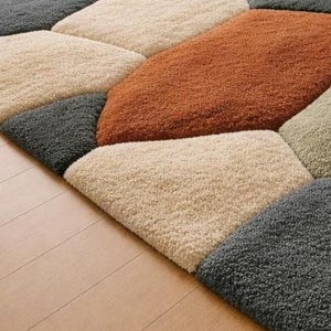 May include: A close-up of a geometric rug with a variety of colors. The rug features a pattern of irregular shapes in shades of beige, gray, and rust. The rug is made of a soft, textured material and is placed on a light-colored wooden floor.