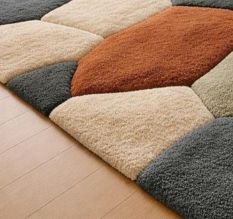May include: A close-up of a geometric rug with a variety of colors. The rug features a pattern of irregular shapes in shades of beige, gray, and rust. The rug is made of a soft, textured material and is placed on a light-colored wooden floor.