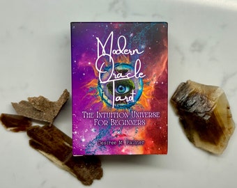 以下が含まれることがあります： 紫と青の銀河のデザインのModern Oracle Tarotカードデッキ。ボックスには「Modern Oracle Tarot」と「The Intuition Universe for Beginners」の文字が書かれています。装飾的な石がデッキの周りに配置されています。