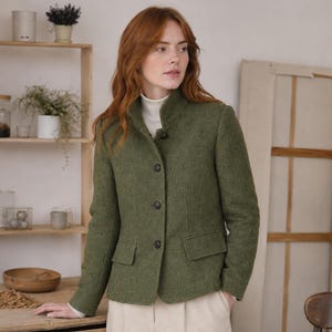 Könnte beinhalten: Grüne Tweedjacke mit Stehkragen und Knopfverschluss. Die Jacke hat zwei Vordertaschen und wird mit beigefarbenen Hosen getragen. Darunter wird ein weißer Rollkragenpullover getragen.