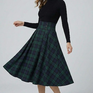 Könnte beinhalten: Ein schwarzer Rollkragenpullover und ein hoch taillierter, ausgestellter karierter Rock in Grün und Marineblau. Der Rock hat einen geknöpften Bund. Das Outfit wird durch schwarze Absätze ergänzt. Der Rock ist etwa 76 cm lang.
